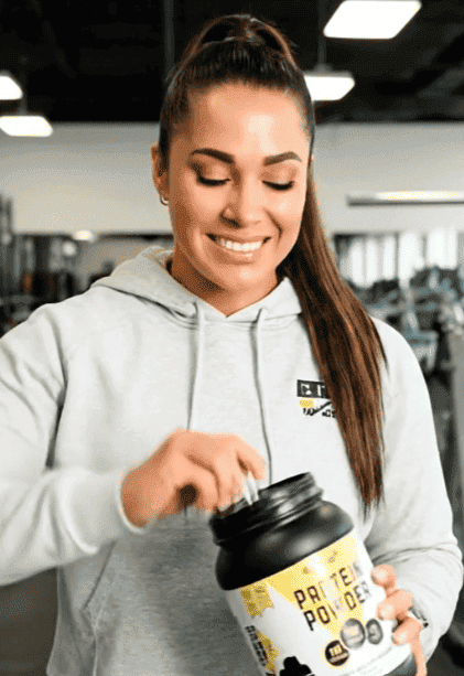 A woman in a gym holding EliteCore chocolate protein powder to make a post-workout protein drink.